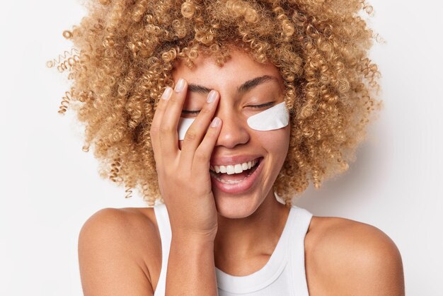 Una joven feliz y positiva con el pelo rizado y tupido hace que la palma de la cara se ría alegremente aplica parches de belleza debajo de los ojos para el tratamiento de la piel vestida con una camiseta informal aislada sobre fondo blanco