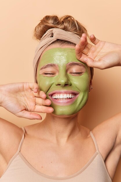 Una joven feliz que toca la cara suavemente aplica una máscara de belleza verde para el cuidado de la piel mantiene los ojos cerrados usa una camiseta con banda para la cabeza se somete a tratamientos para el cuidado de la piel aislados sobre un fondo beige. concepto de bienestar