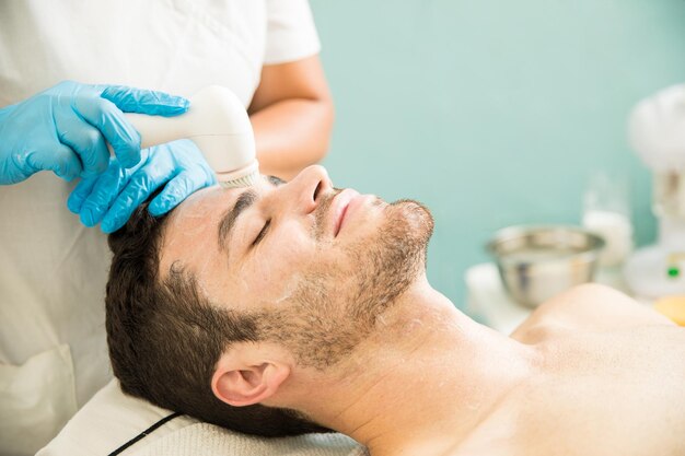 Un joven feliz recibiendo una exfoliación facial y un exfoliante con un cepillo en un spa de salud y belleza