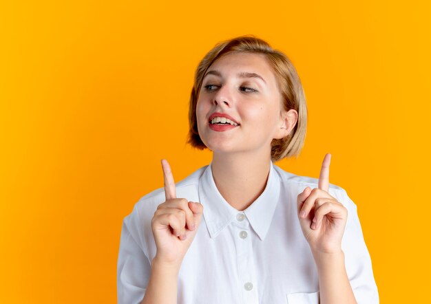 Joven feliz rubia rusa apunta hacia arriba aislado sobre fondo naranja con espacio de copia