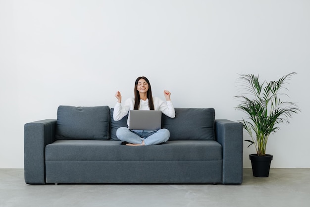 Una joven feliz sentada en el sofá de su casa triunfante recibe un correo electrónico agradable o lee buenas noticias en una computadora portátil en línea