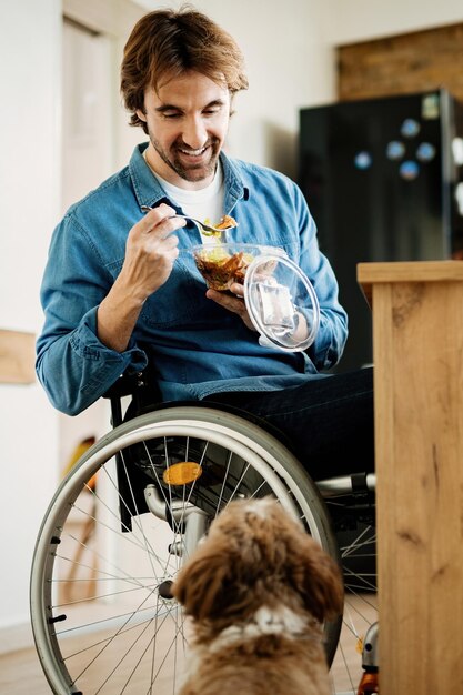 Foto gratuita joven feliz en silla de ruedas comiendo mientras está con su perro en casa.