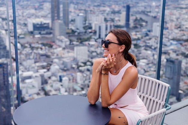 Joven feliz sonriente mujer caucásica viajero en vestido ajustado y gafas de sol en piso alto en Bangkok Elegante mujer explorando increíbles vistas de la gran ciudad