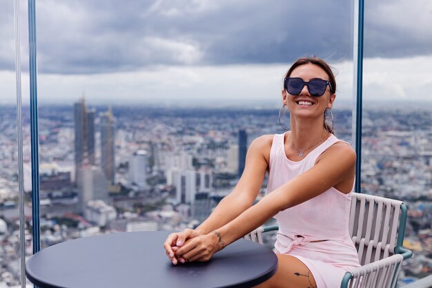 Joven feliz sonriente mujer caucásica viajero en vestido ajustado y gafas de sol en piso alto en Bangkok Elegante mujer explorando increíbles vistas de la gran ciudad