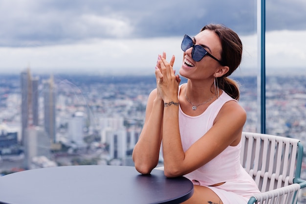 Joven feliz sonriente mujer caucásica viajero en vestido ajustado y gafas de sol en piso alto en Bangkok Elegante mujer explorando increíbles vistas de la gran ciudad