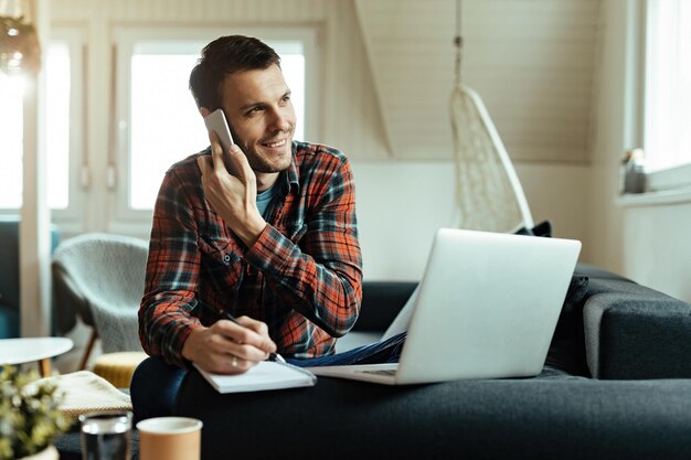 Joven feliz tomando notas y comunicándose por teléfono celular mientras usa una computadora en la sala de estar