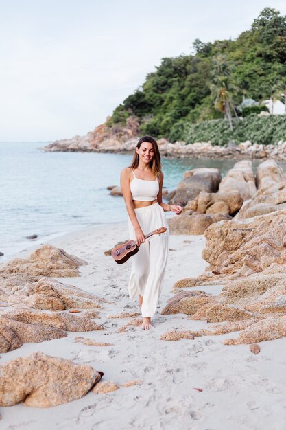 Joven feliz tranquila mujer caucásica con ukelele en la playa rocosa tropical al atardecer