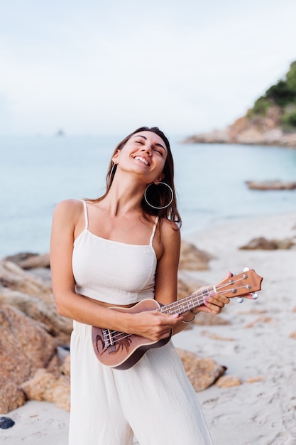 Joven feliz tranquila mujer caucásica con ukelele en la playa rocosa tropical al atardecer