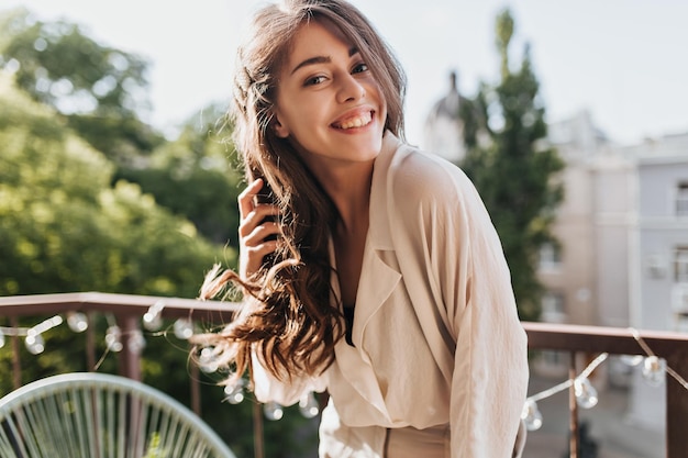 Joven felizmente posando en la terraza Retrato de mujer morena rizada en blusa beige sonriendo ampliamente y relajándose en el balcón