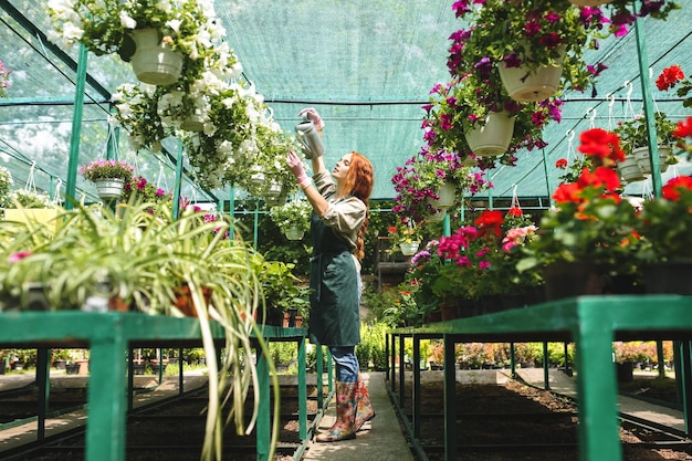 Joven florista bonita en delantal de pie y regando soñadoramente hermosas flores mientras trabaja en un gran invernadero