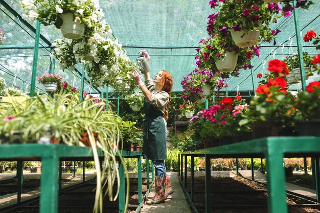 Joven florista bonita en delantal de pie y regando soñadoramente hermosas flores mientras trabaja en un gran invernadero