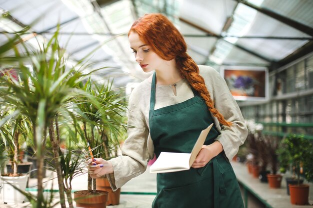 Joven florista en delantal de pie con bloc de notas y lápiz en las manos mientras trabaja con plantas en invernadero