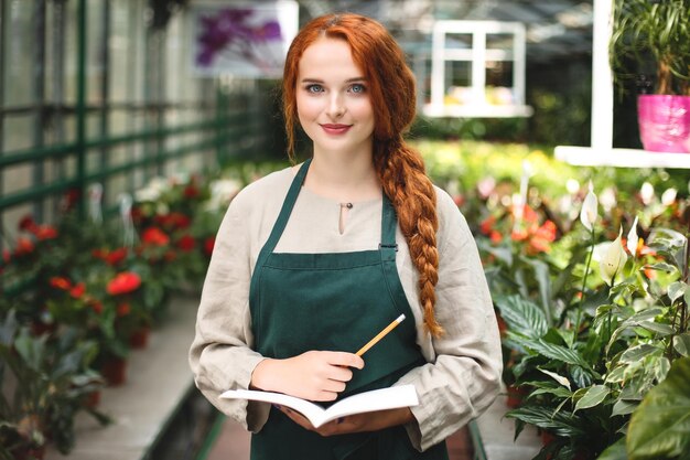 Joven florista sonriente en delantal de pie con bloc de notas y lápiz en las manos y felizmente mirando a la cámara mientras trabaja en invernadero