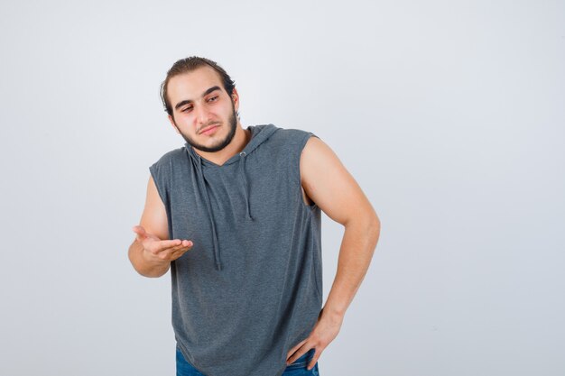 Joven en forma masculina extendiendo la palma en sudadera con capucha sin mangas y luciendo confiado. vista frontal.