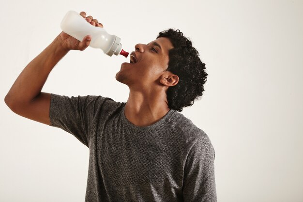 Joven en forma sonriente negro bebiendo agua de una botella deportiva, con la boca abierta, aislado en blanco