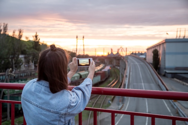 Foto gratuita una joven fotografía una hermosa puesta de sol desde un puente con su teléfono.