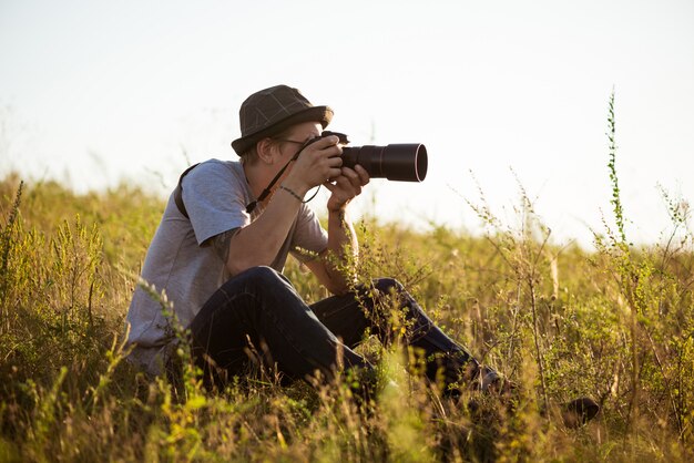 Joven fotógrafo masculino con sombrero tomando foto, sentado en el campo