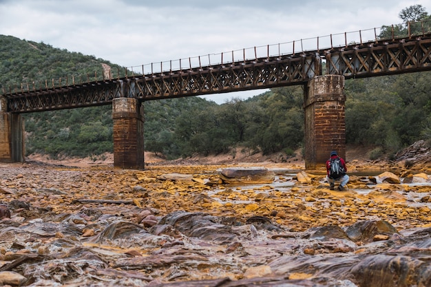 Foto gratuita joven fotógrafo con mochila y gorra fotografiando un viejo puente de hierro sobre el río tinto.