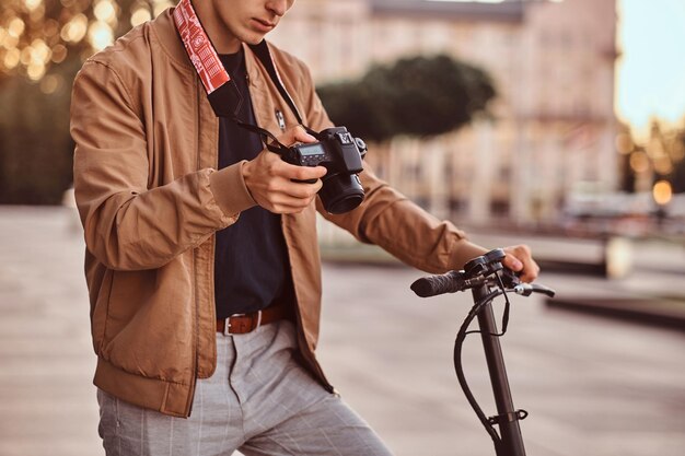 Joven fotógrafo pensativo con scooter está viendo fotos en su cámara de fotos digital mientras viaja.