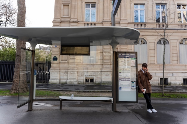 Joven francés esperando el autobús en la estación y hablando por teléfono inteligente