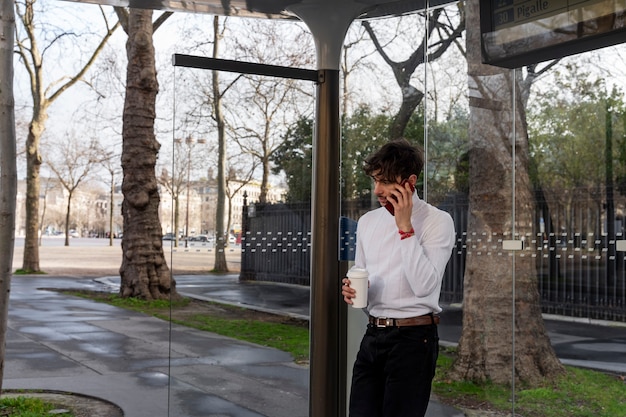 Joven francés esperando el autobús en la estación y hablando por teléfono inteligente