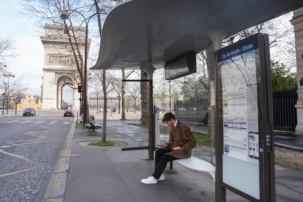 Joven francés esperando en la estación el autobús y usando su teléfono inteligente