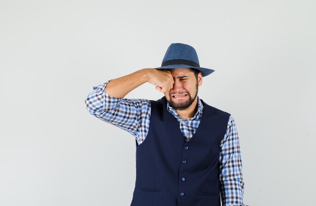 Joven frotándose los ojos mientras llora en camisa, chaleco, sombrero y mirando ofendido.