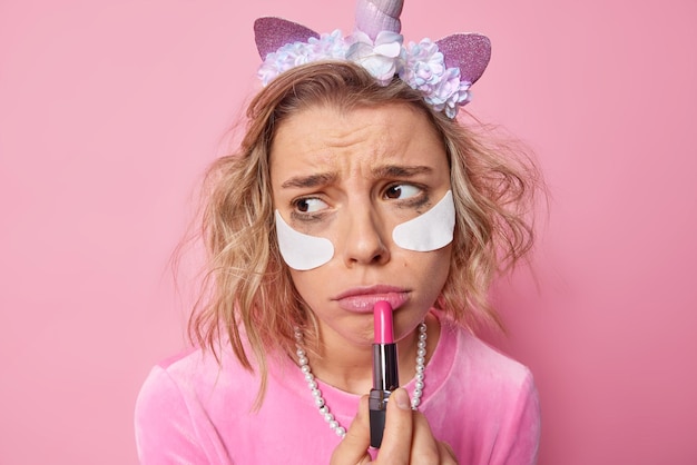 Foto gratuita la joven frustrada se aplica el lápiz labial y se ve tristemente lejos, se ha estropeado el maquillaje, se pone parches blancos debajo de los ojos, se peina, usa una diadema de unicornio y posa contra un fondo rosa. concepto de belleza.