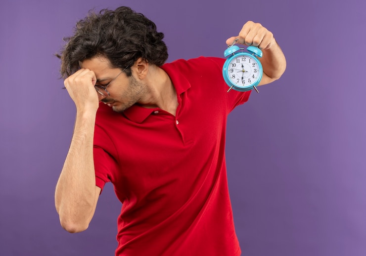 Foto gratuito joven frustrado en camisa roja con gafas ópticas sostiene el reloj y pone la mano en la cara aislada en la pared violeta