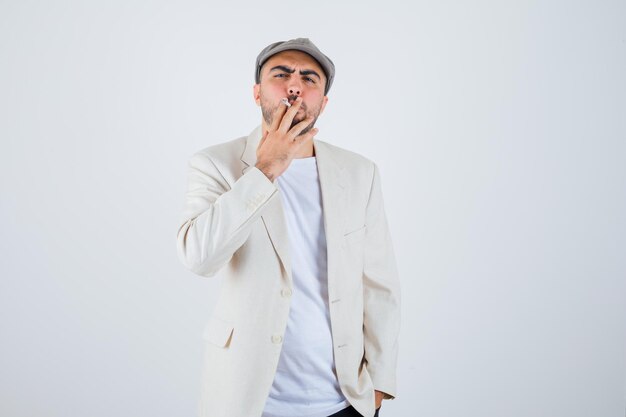 Joven fumando cigarrillos en camiseta blanca, chaqueta y gorra gris y mirando serio