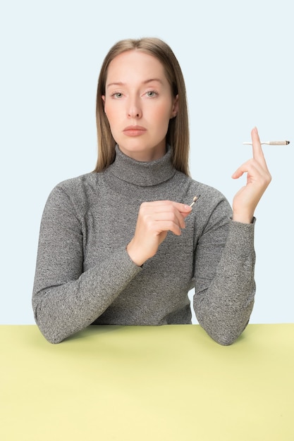 La joven fumando cigarrillos mientras está sentado a la mesa en el estudio. Colores de moda. El retrato de niña caucásica en estilo minimalista con espacio de copia