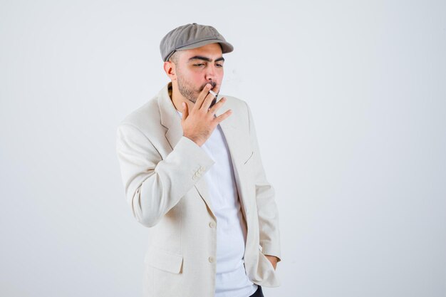 Joven fumando cigarrillos mientras sostiene las manos en la cintura con camiseta blanca, chaqueta y gorra gris y mirando serio