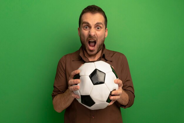 Joven furioso sosteniendo un balón de fútbol mirando al frente gritando aislado en la pared verde
