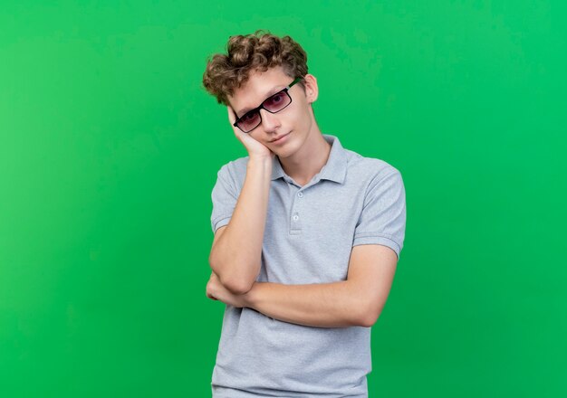 Joven de gafas negras con camisa de polo gris con la cabeza inclinada sobre su brazo esperando sobre verde