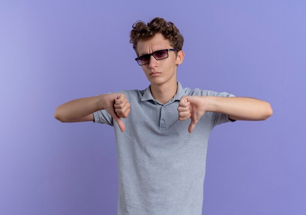 Joven de gafas negras con camisa de polo gris con cara seria apuntando con los dedos hacia abajo de pie sobre la pared azul