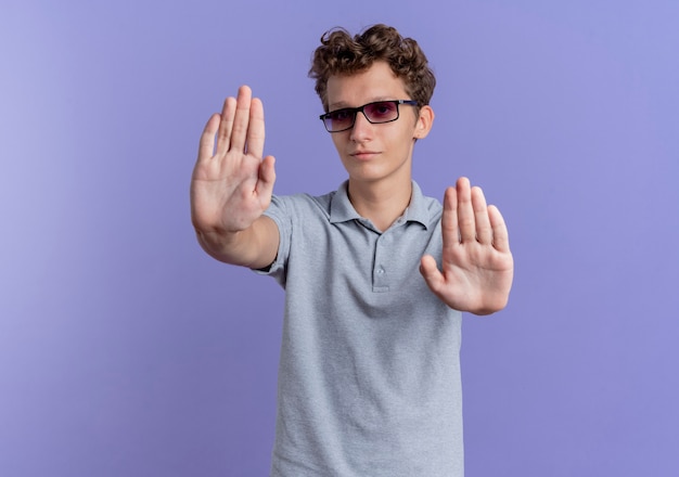 Joven de gafas negras con camisa de polo gris con cara seria haciendo gesto de parada con las manos de pie sobre la pared azul