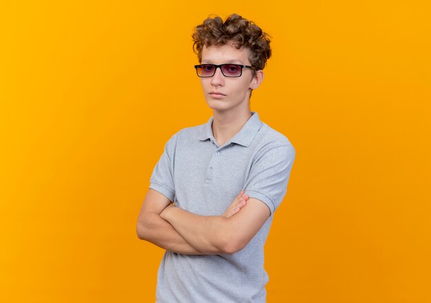 Joven de gafas negras con camisa de polo gris con cara seria con las manos cruzadas sobre el pecho de pie sobre la pared naranja