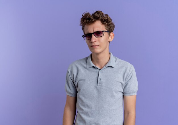 Joven de gafas negras con camisa de polo gris con el ceño fruncido de pie sobre la pared azul