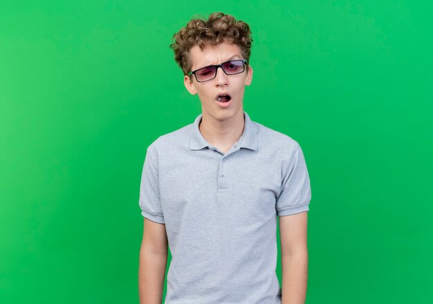 Joven de gafas negras con camisa de polo gris con expresión escéptica de pie sobre la pared verde