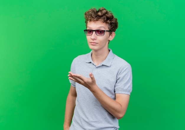 Joven de gafas negras con camisa de polo gris gesticulando con el brazo sobre verde