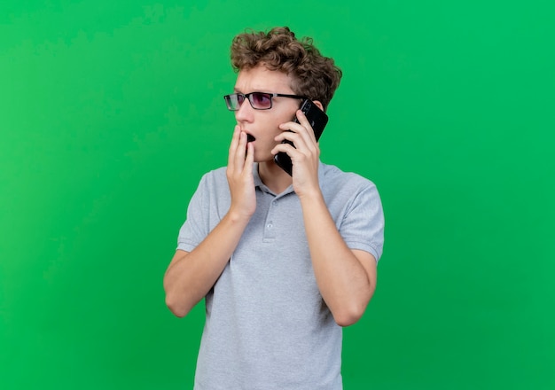 Joven de gafas negras con camisa de polo gris hablando por teléfono móvil asombrado y sorprendido sobre verde