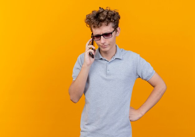 Joven de gafas negras con camisa de polo gris hablando por teléfono móvil confundido y disgustado por naranja