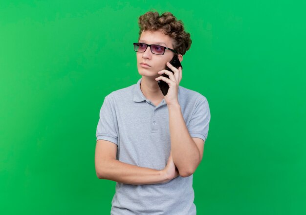 Joven de gafas negras con camisa de polo gris hablando por teléfono móvil mirando con cara seria sobre verde