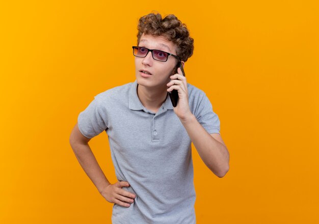 Joven de gafas negras con camisa de polo gris hablando por teléfono móvil mirando sorprendido de pie sobre la pared naranja