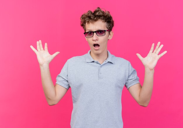 Joven de gafas negras con camisa de polo gris levantando las palmas mirando sorprendido de pie sobre la pared rosa