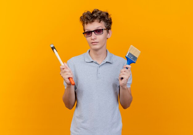 Joven de gafas negras con camisa de polo gris con martillo y pincel con cara seria sobre naranja