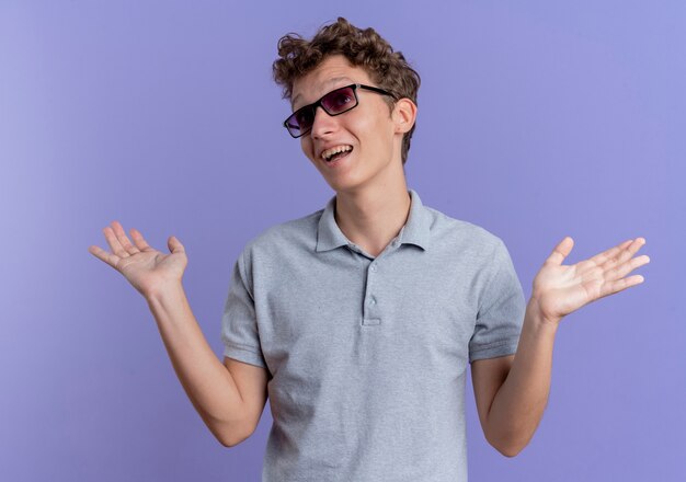 Joven de gafas negras con camisa de polo gris mirando confundido e incierto levantando las manos sin respuesta de pie sobre la pared azul