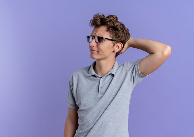 Joven de gafas negras con camisa de polo gris mirando a un lado confundido y muy ansioso de pie sobre la pared azul