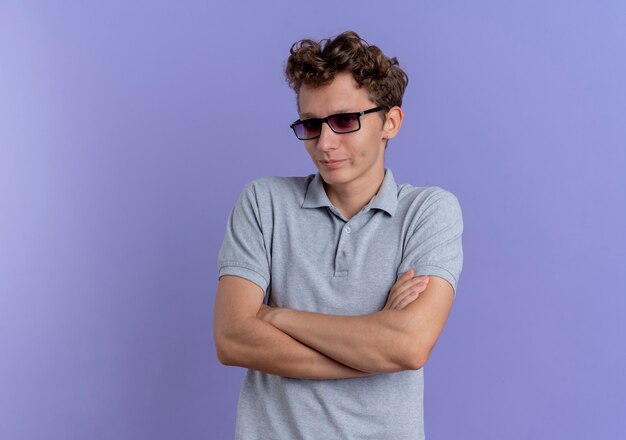 Joven de gafas negras con camisa de polo gris mirando a un lado disgustado con las manos cruzadas sobre el pecho de pie sobre la pared azul