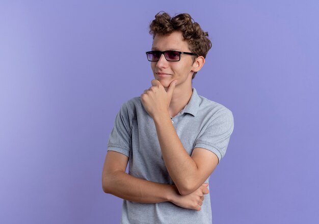 Joven de gafas negras con camisa polo gris mirando a un lado con la mano en la barbilla con expresión pensativa sobre azul